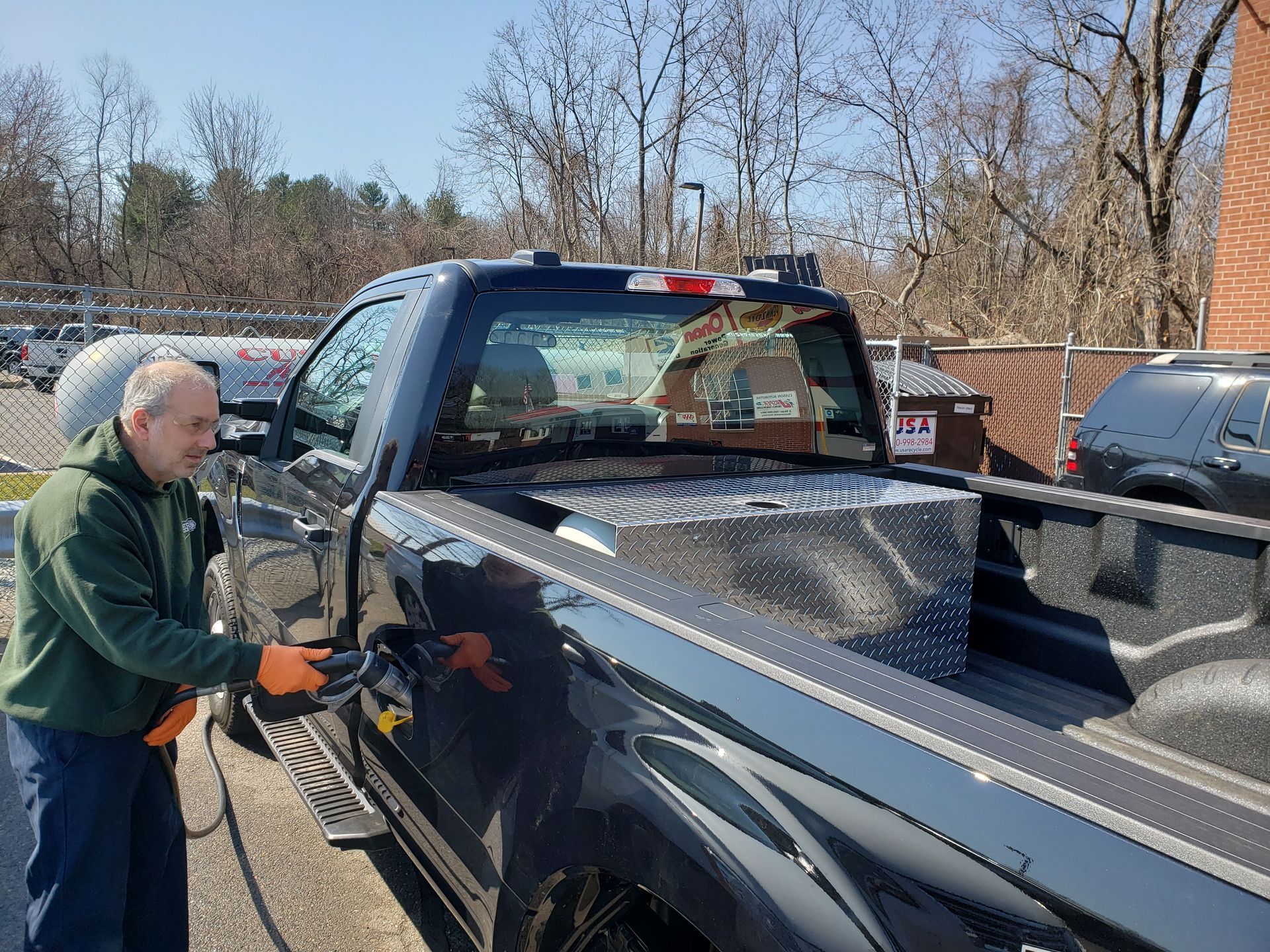 Man fueling a black pickup truck at a gas station with a wooded background