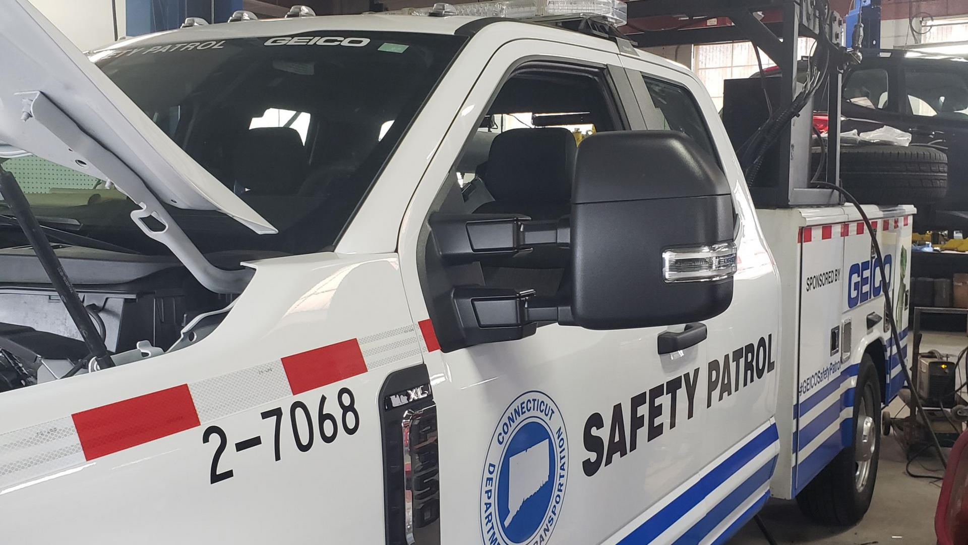 White safety patrol vehicle with open door and raised hood in an auto shop, marked “2-7068” and “SAFETY PATROL”