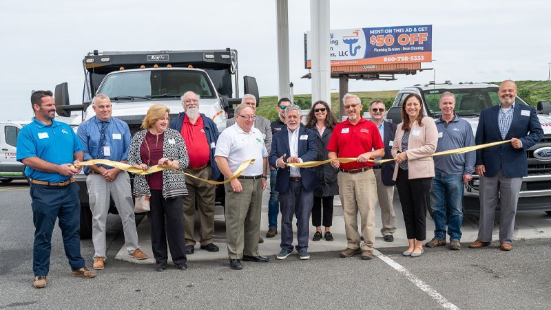 Group of people at a ribbon-cutting ceremony in front of a truck and roadside sign.