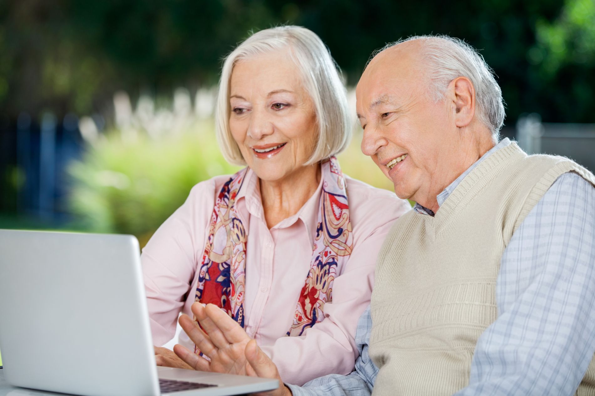 Coppia di anziani sorridenti, che guardano un computer portatile all'aperto. La donna indica, l'uomo sorride.