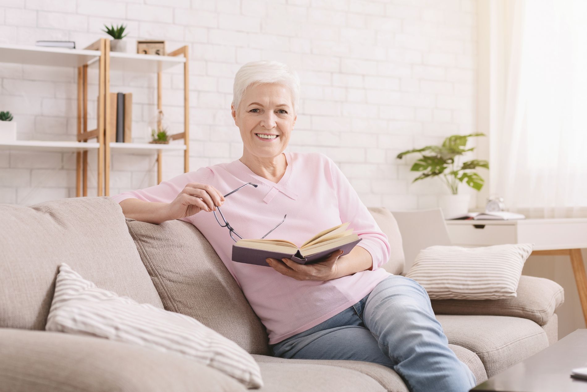 Una donna anziana sorridente con i capelli corti e bianchi è seduta su un divano, con in mano un libro e degli occhiali, in un luminoso soggiorno.