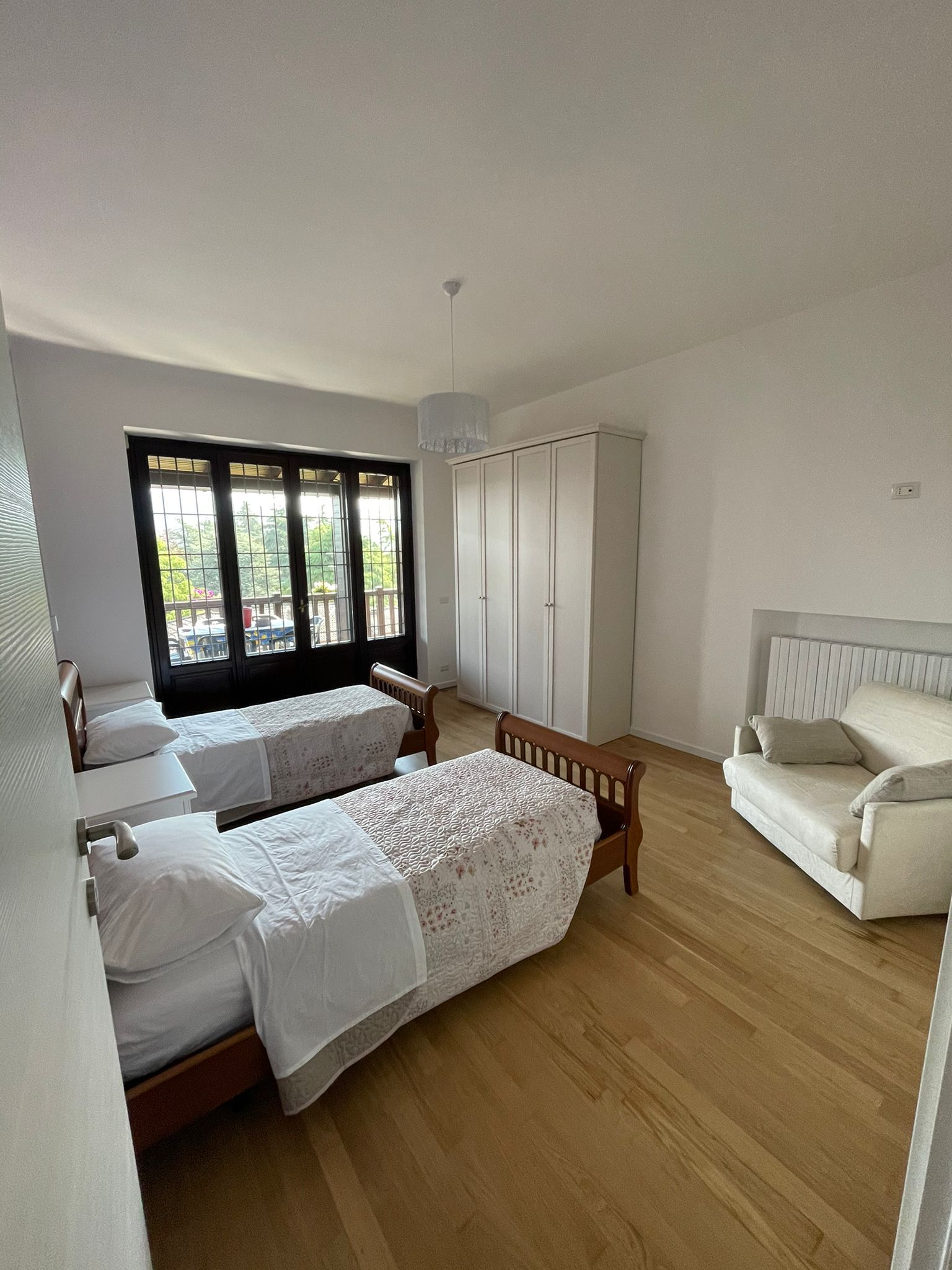 Camera da letto con due letti singoli, armadio bianco e un piccolo divano; ampia finestra e pavimento in legno chiaro.