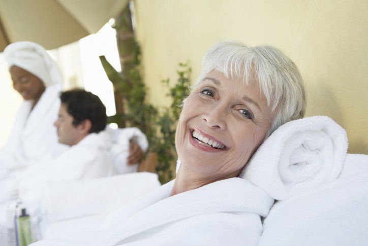 Donna sorridente in accappatoio da spa con un asciugamano dietro la testa; altre due persone sono sfocate sullo sfondo.