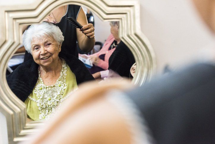 Una donna anziana sorride mentre si fa acconciare i capelli in un salone, riflessa in uno specchio.