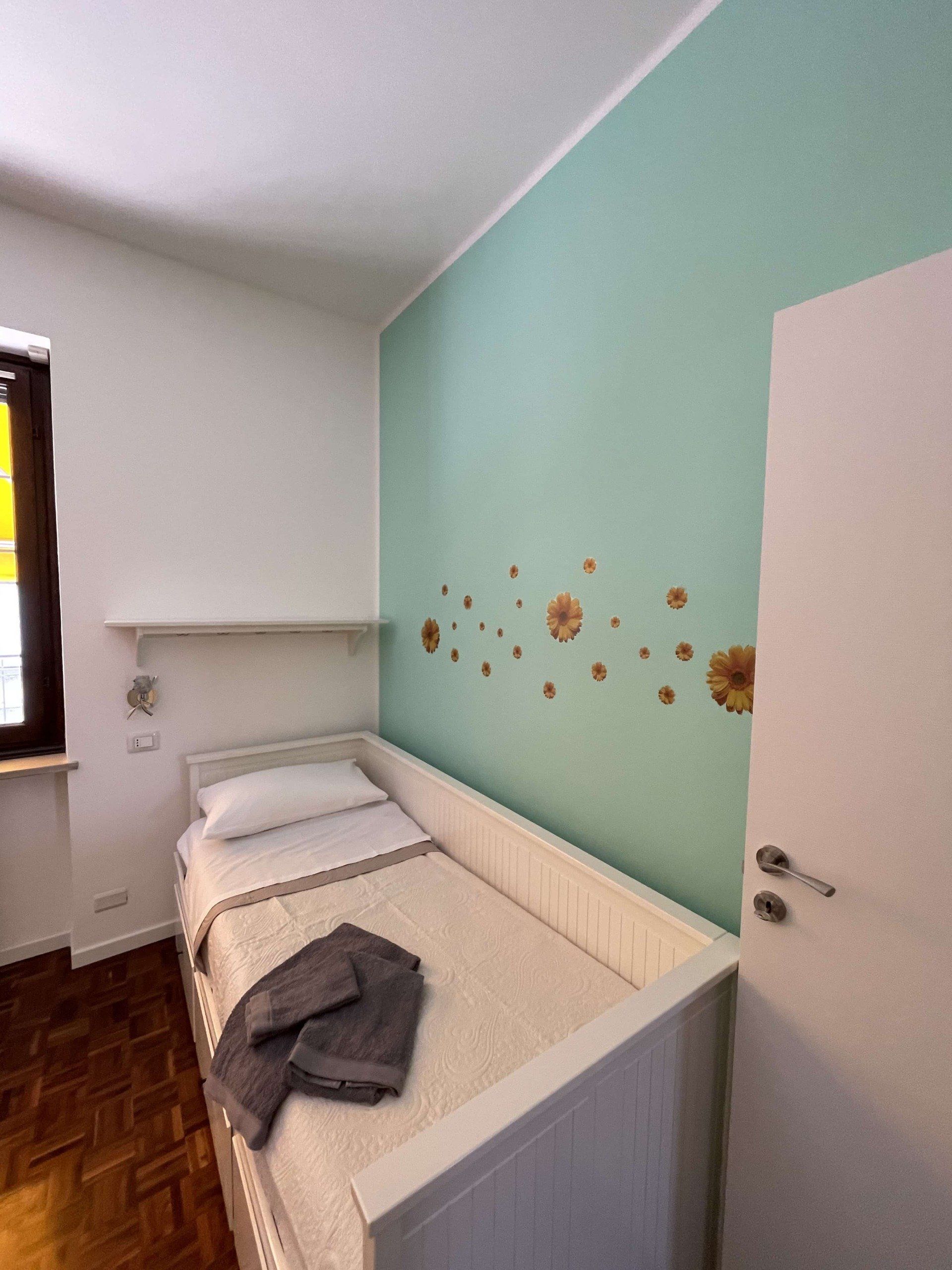 Piccola camera da letto con letto bianco, parete turchese con decorazioni floreali e porta bianca.