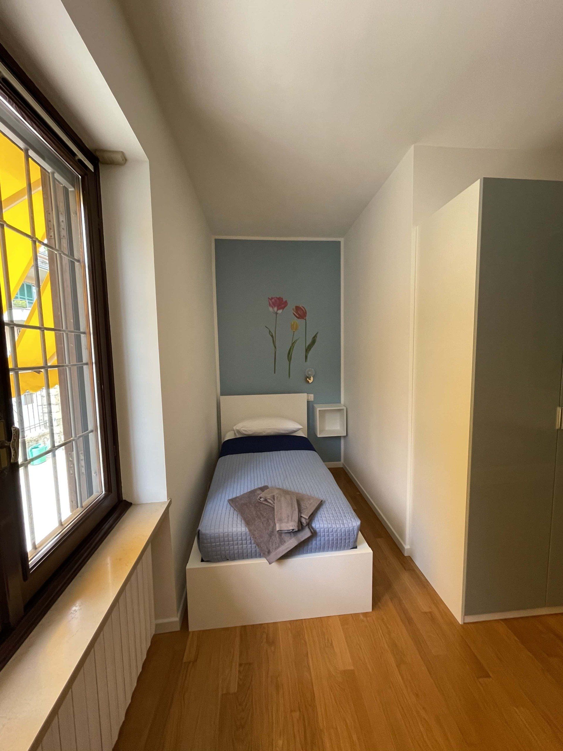 Camera da letto stretta con un letto singolo, pavimenti in legno e una finestra sulla sinistra.
