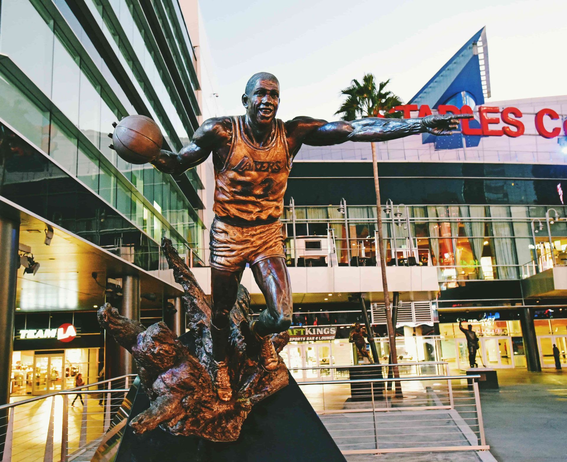 Bronze Los Angeles Lakers statue outside the Staples Center area in downtown Los Angeles, with arena signage, palm trees, and city lights in the background.