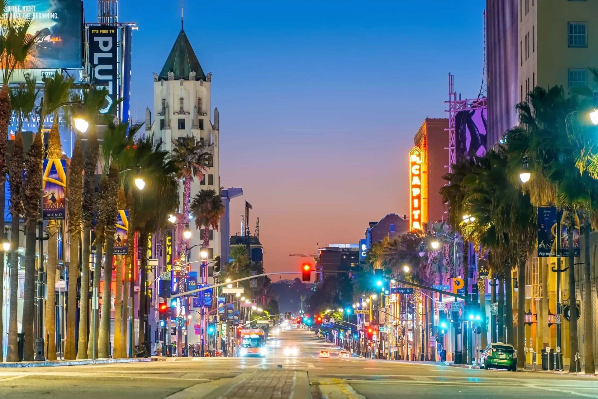 Downtown Los Angeles nightlife along a palm-lined street with illuminated theater signs, city lights, and traffic at dusk.