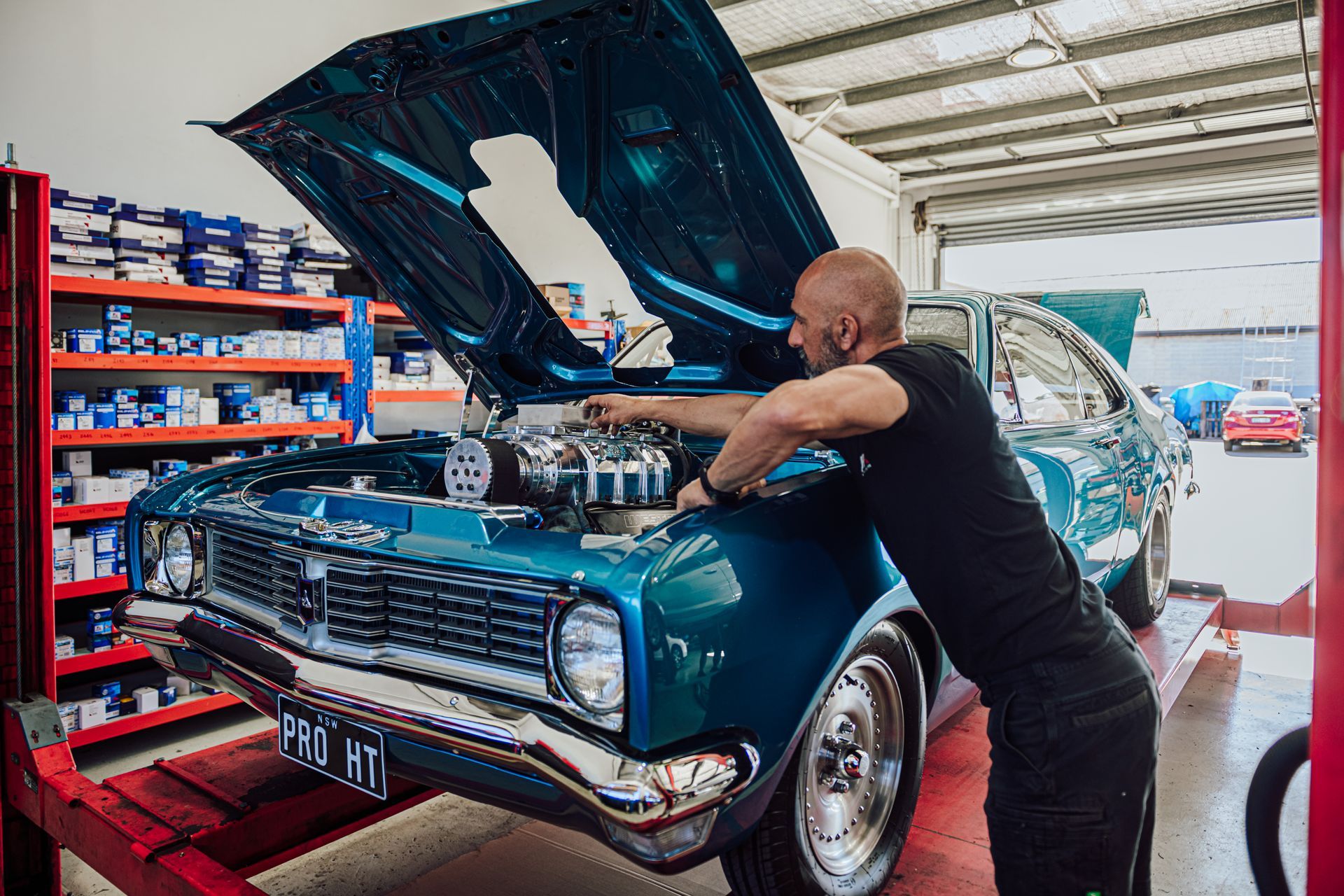 A mechanic working on the open hood of a teal classic car inside a garage.