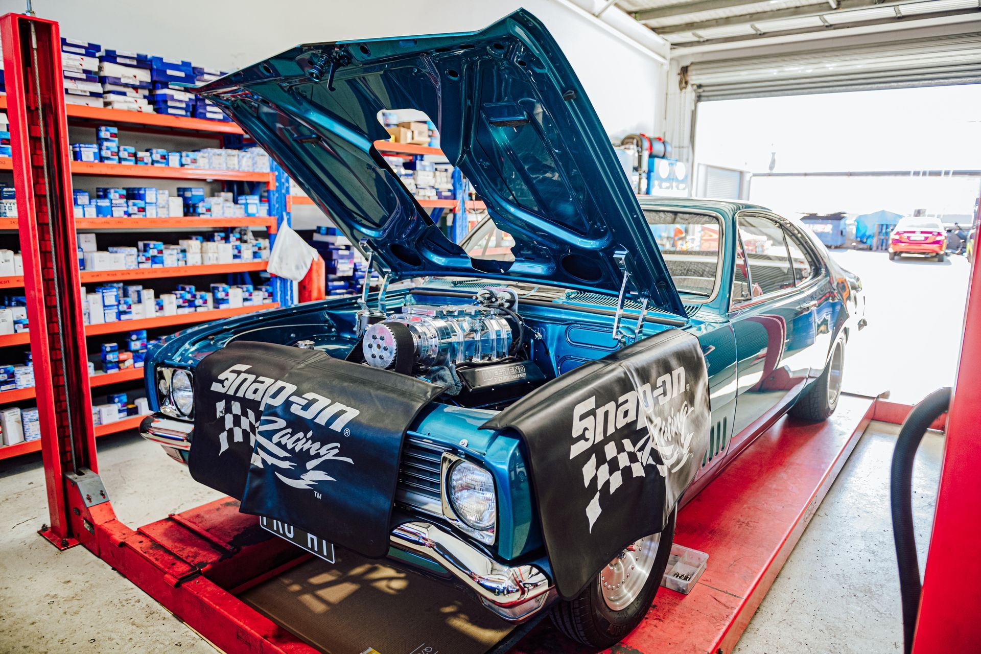 Blue classic car on a lift in a garage, hood open, engine visible. Snap-on Racing cover on the fenders.