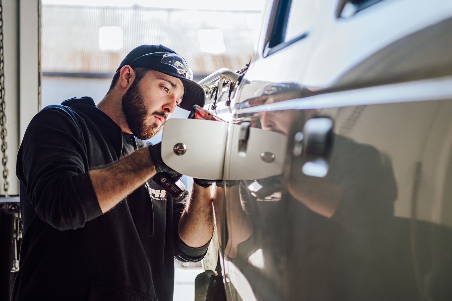 Mechanic in black gloves and hat installing a white side mirror on a white car in a garage.