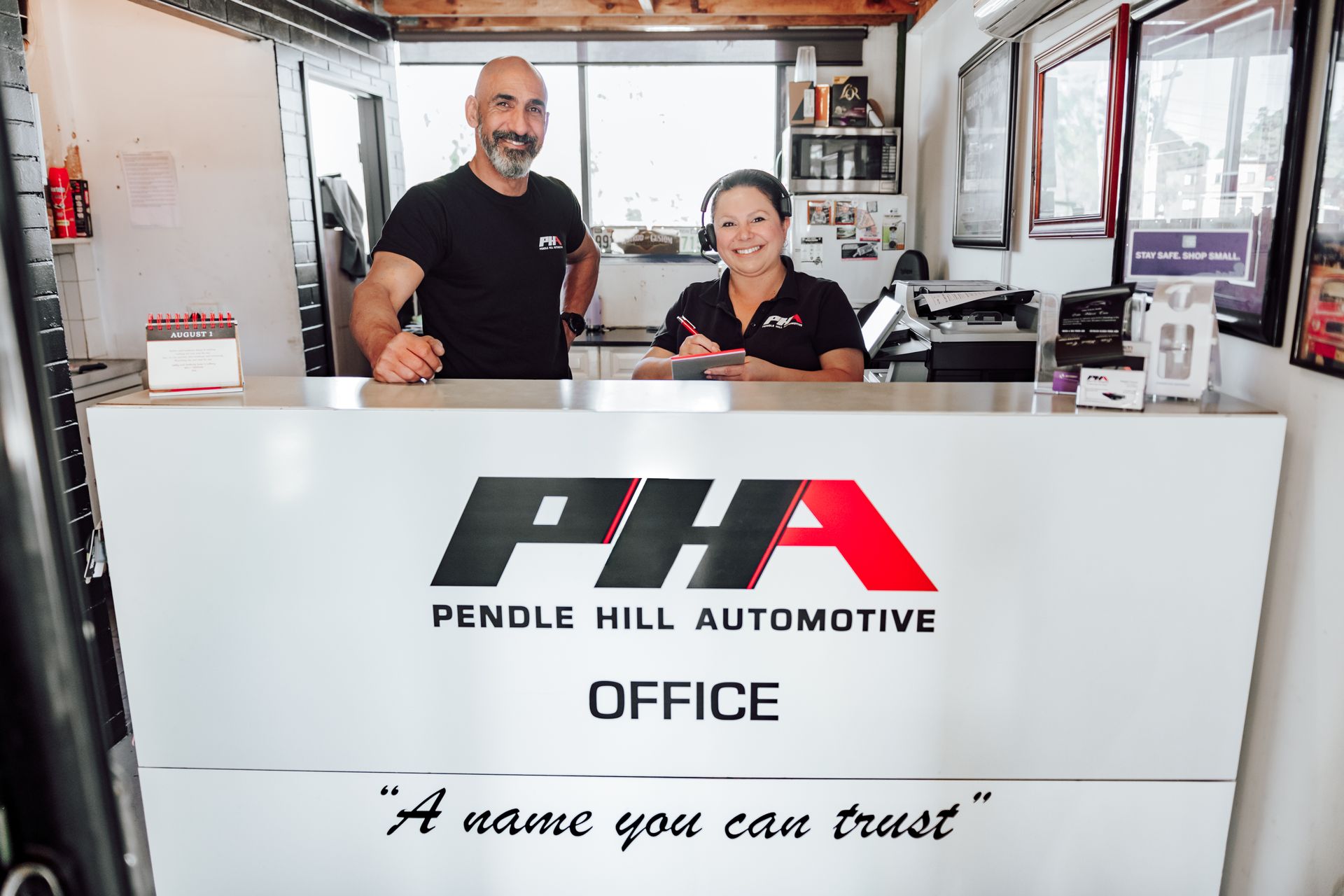 Two people at the reception desk of Pendle Hill Automotive office. Man and woman smile, logo visible.