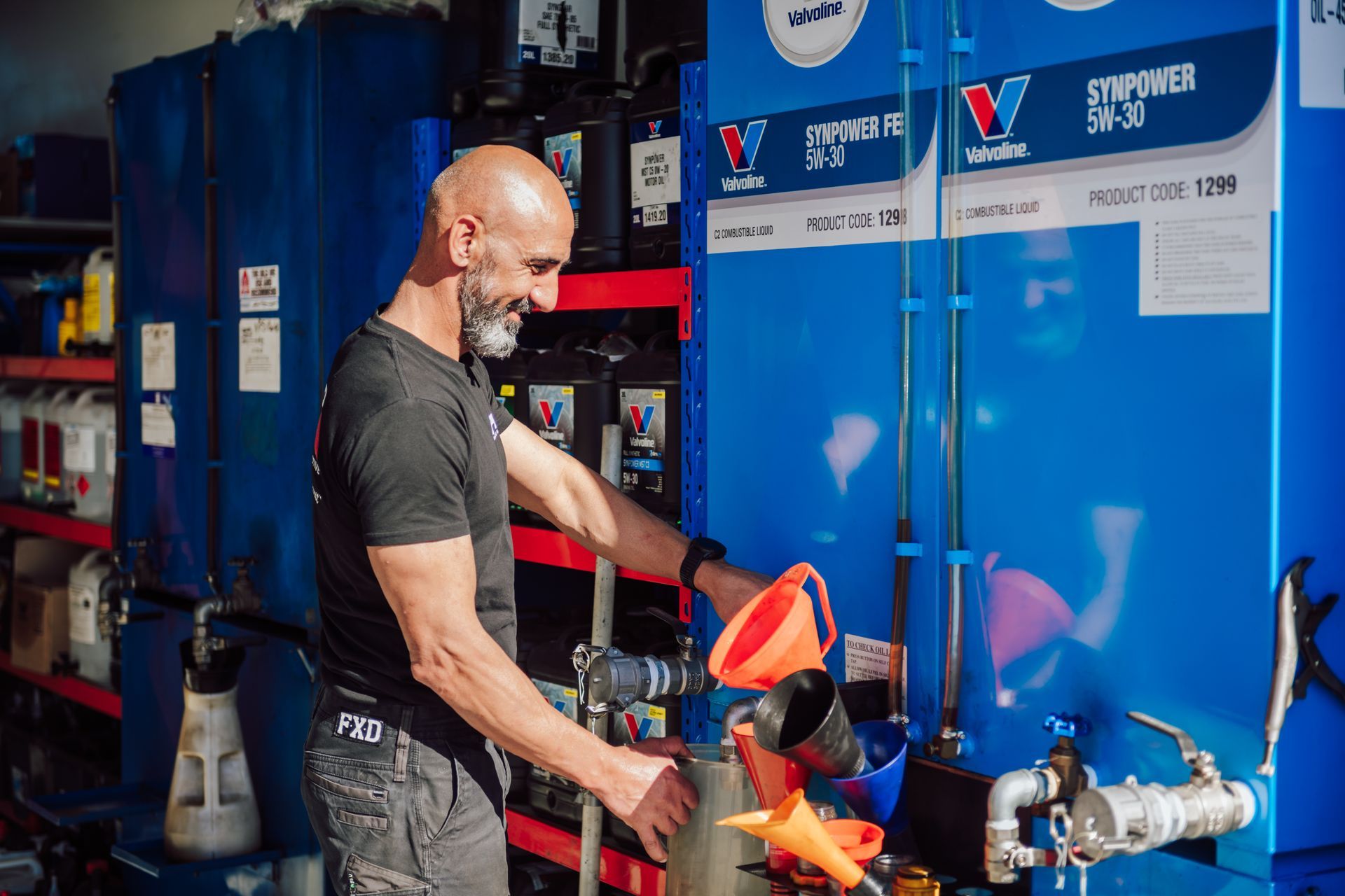 Man pouring oil from a blue tank with a funnel in a shop setting.