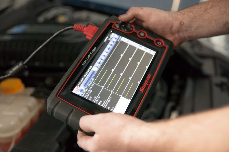 Hands holding a Snap-on diagnostic tool, connected to a car engine, displaying waveform data on its screen.
