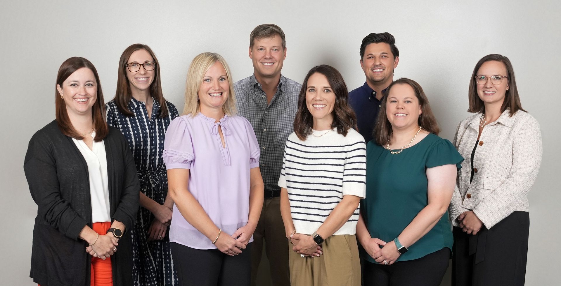 Seven coworkers standing in a row against a gray studio backdrop, smiling at the camera.
