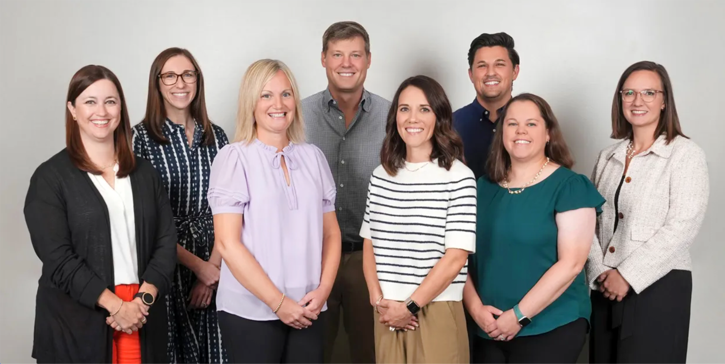 Seven people standing together in a studio, smiling at the camera against a light background.
