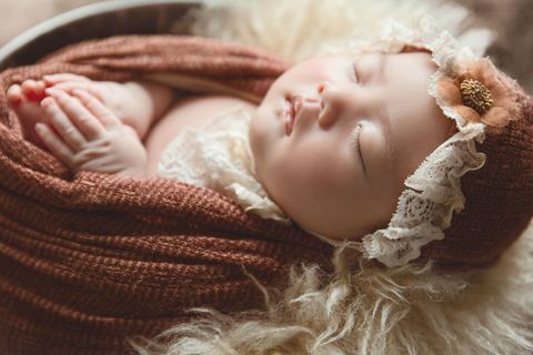 Sleeping baby swaddled in a brown knit blanket with lace bonnet on a soft cream fur background