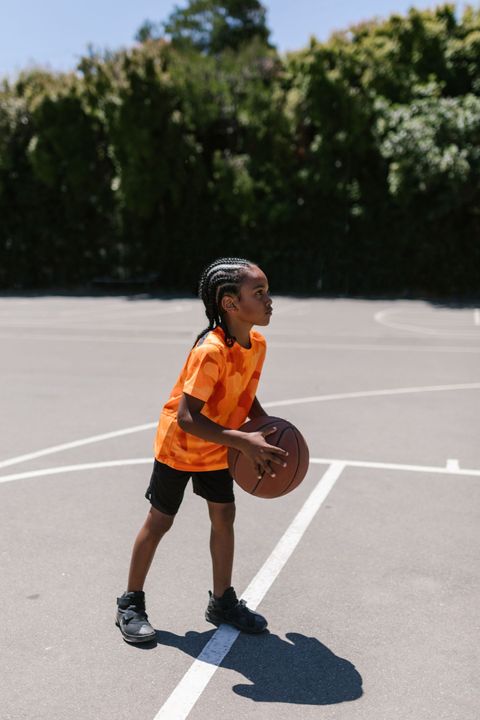 Child dribbling a basketball on an outdoor court, wearing an orange jersey and black shorts.