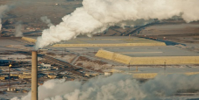 Smokestack billowing white smoke into the air over an industrial area and distant buildings.