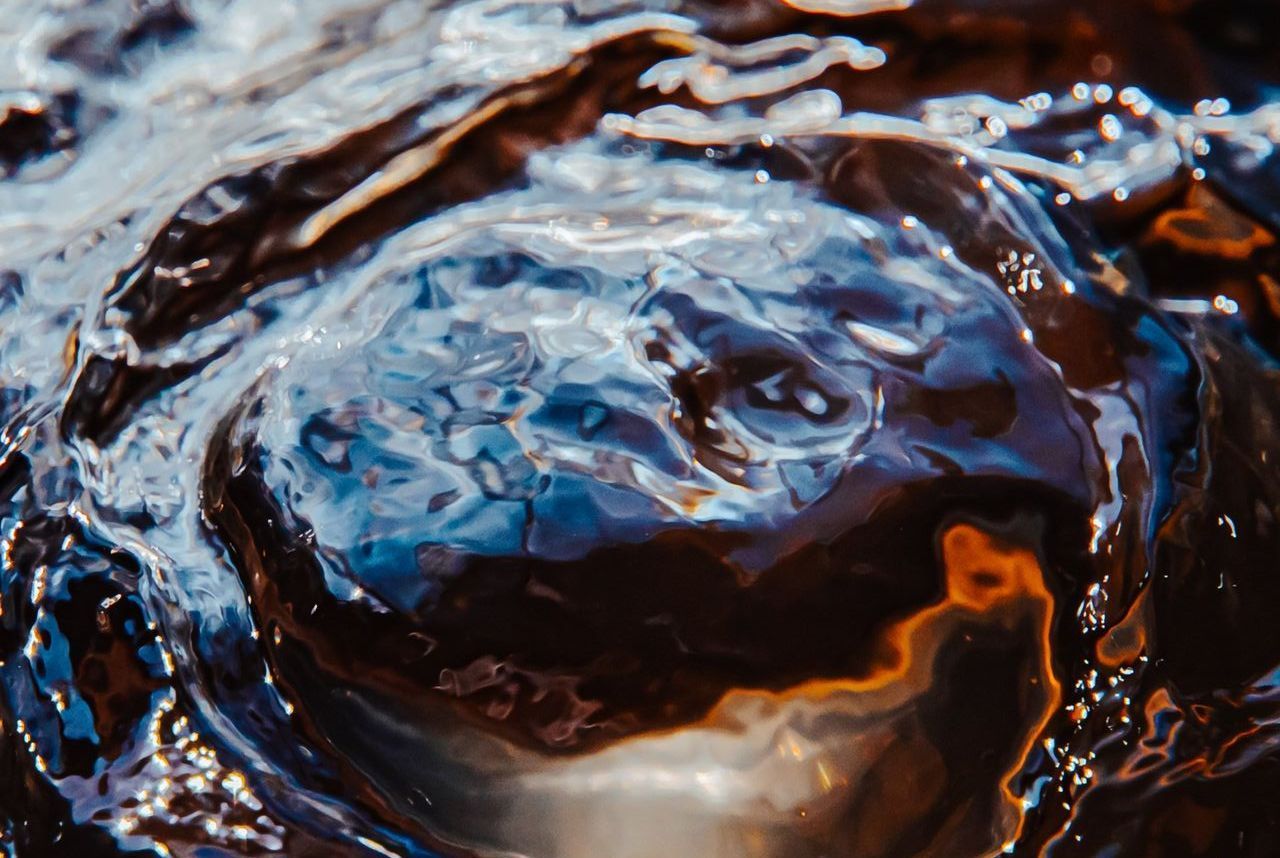 Close-up of rippling water with dark brown and blue reflections, highlighted by orange and white light.