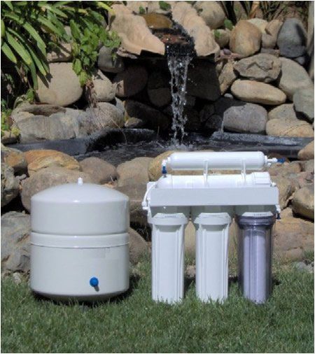 Water filter system with a white tank on grass in front of a waterfall feature.