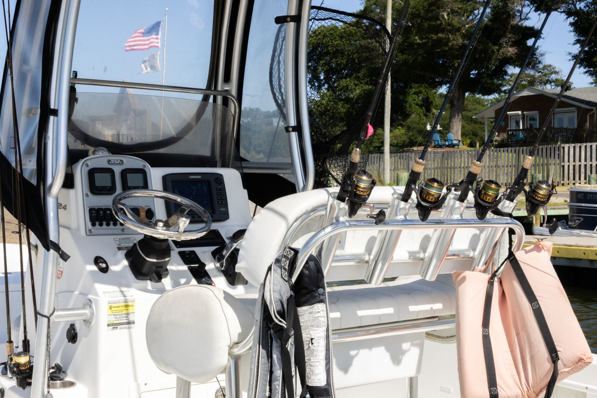 Boat cockpit with white seats, steering wheel, and flag, overlooking a marina with buildings and trees