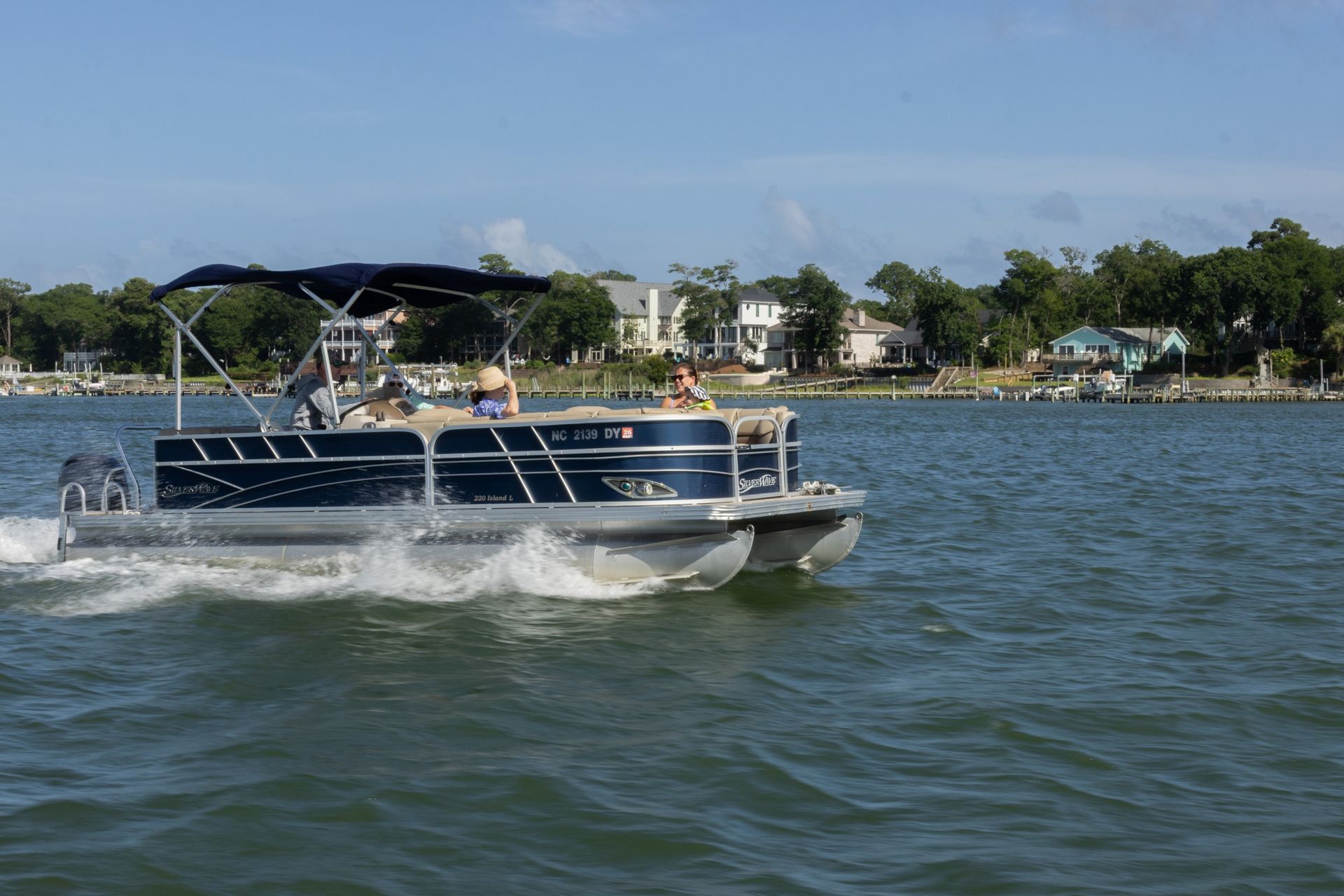 Pontoon boat cruising on a lake, splashing water near a shoreline with houses and trees