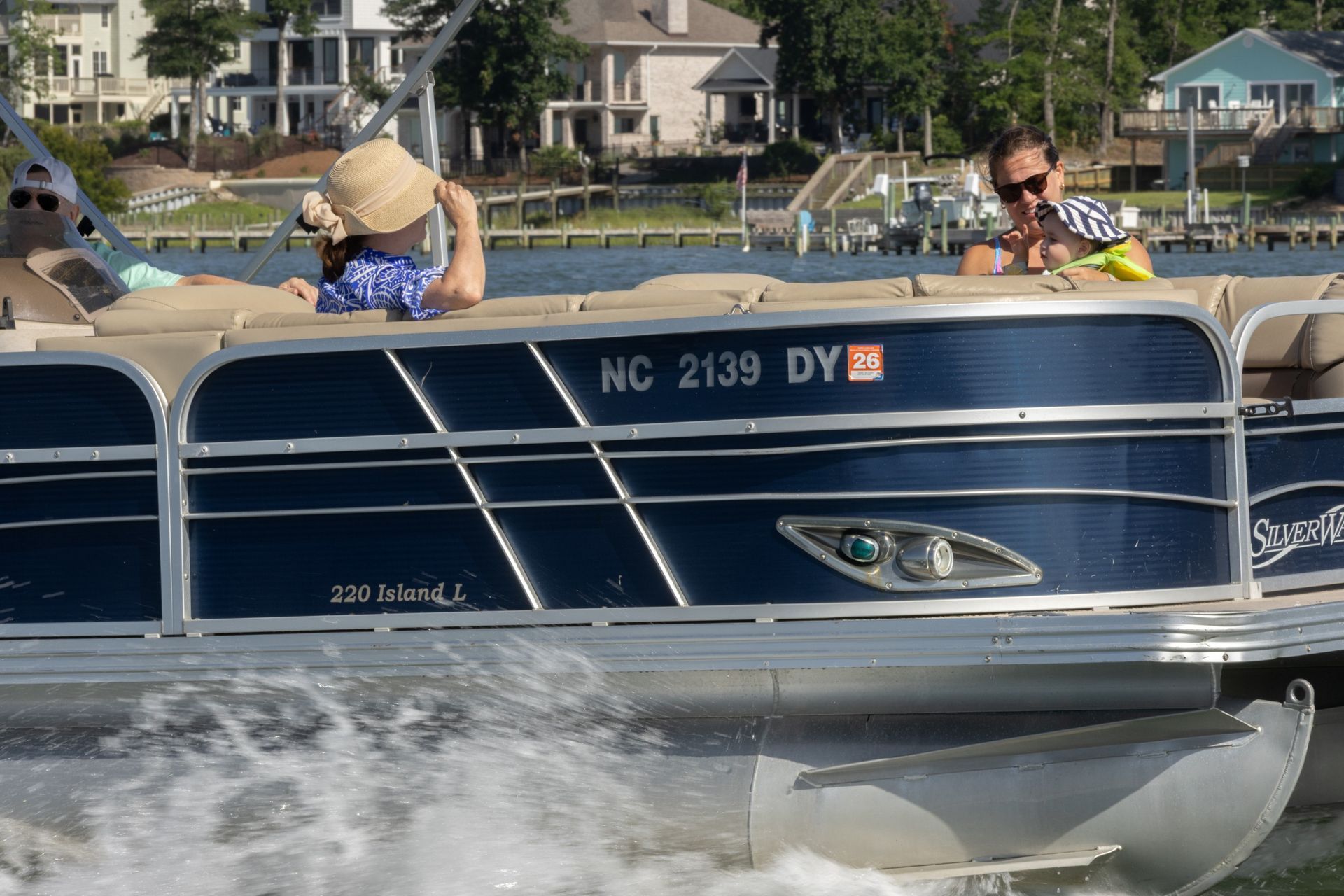 Blue pontoon boat on a lake with two people lounging, houses and trees in the background.