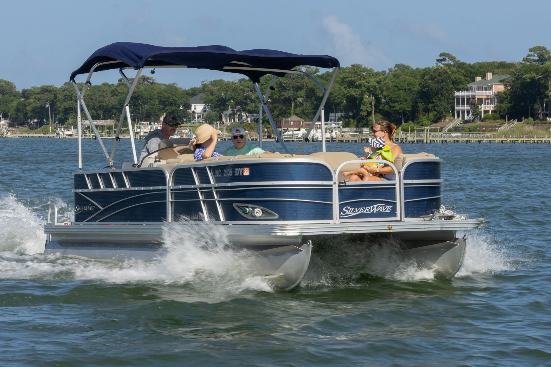Pontoon boat cruising on a lake, with passengers under a blue canopy and water splashing around it.
