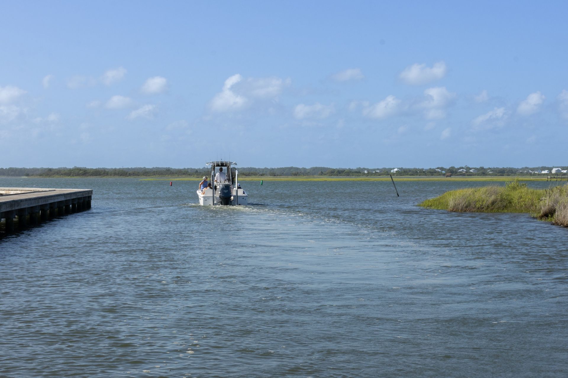Small motorboat on a calm blue inlet under a clear sky, with a dock on the left and marshy shore on the right