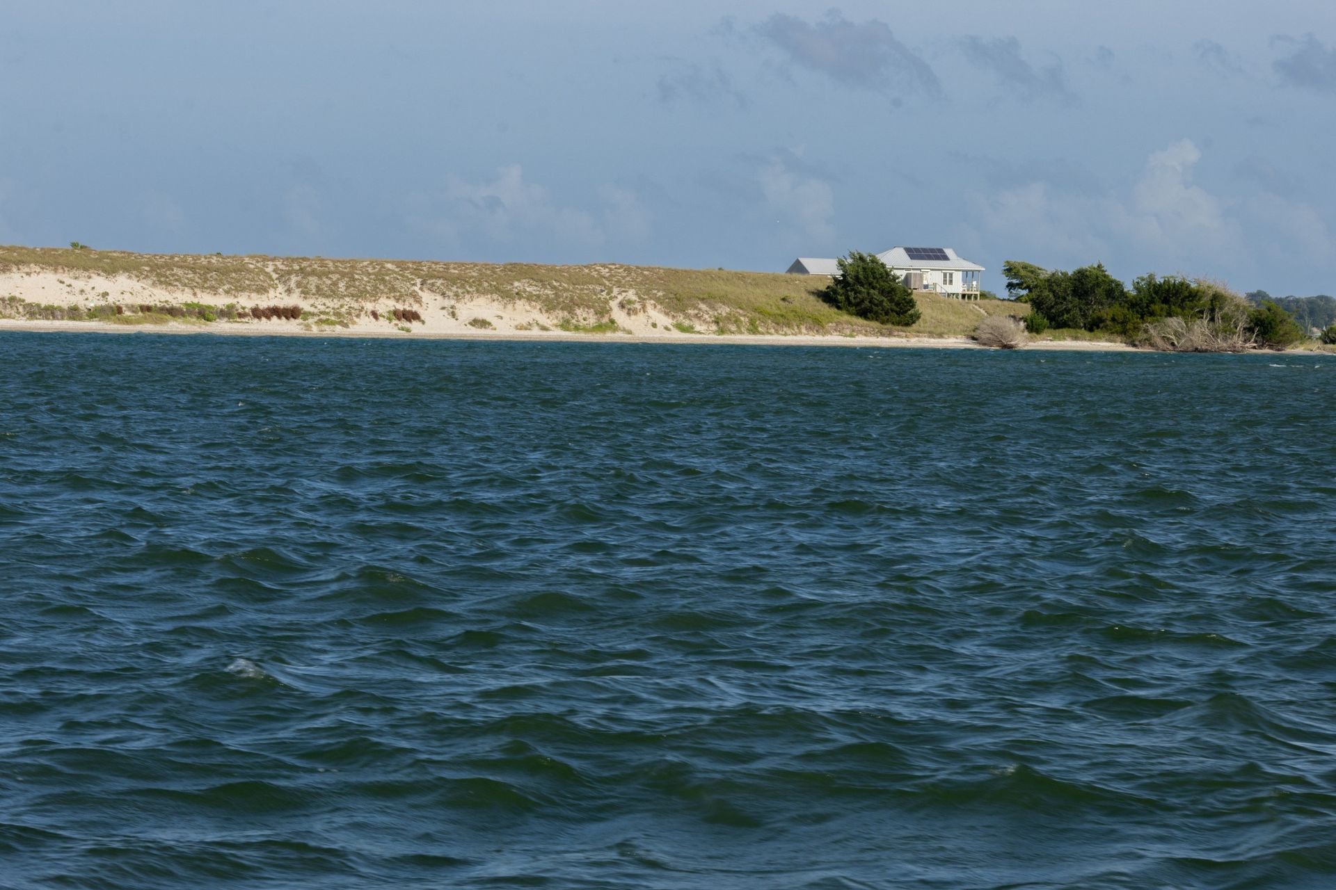 Choppy blue water with a sandy shoreline, small white house, and cloudy sky in the distance