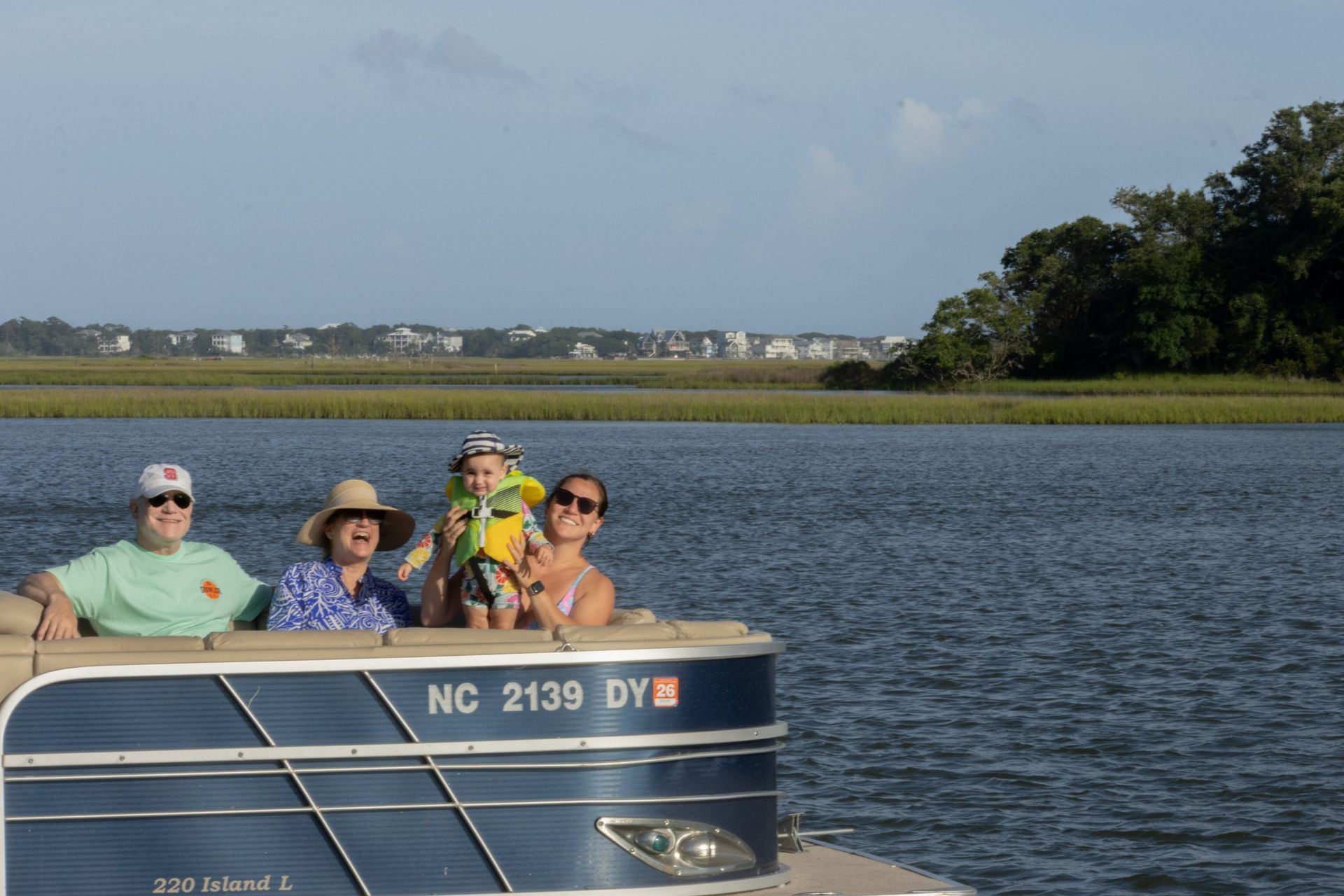 Four people smiling on a blue boat on calm water near a grassy shoreline under a clear sky