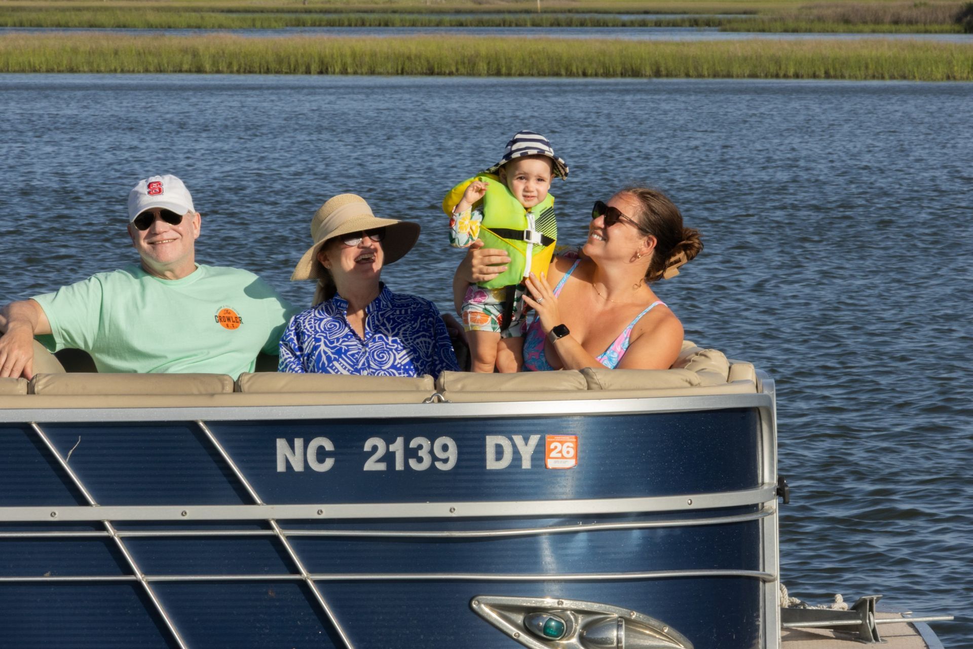 People riding in a pontoon boat on a lake, with blue water and marshland behind them.