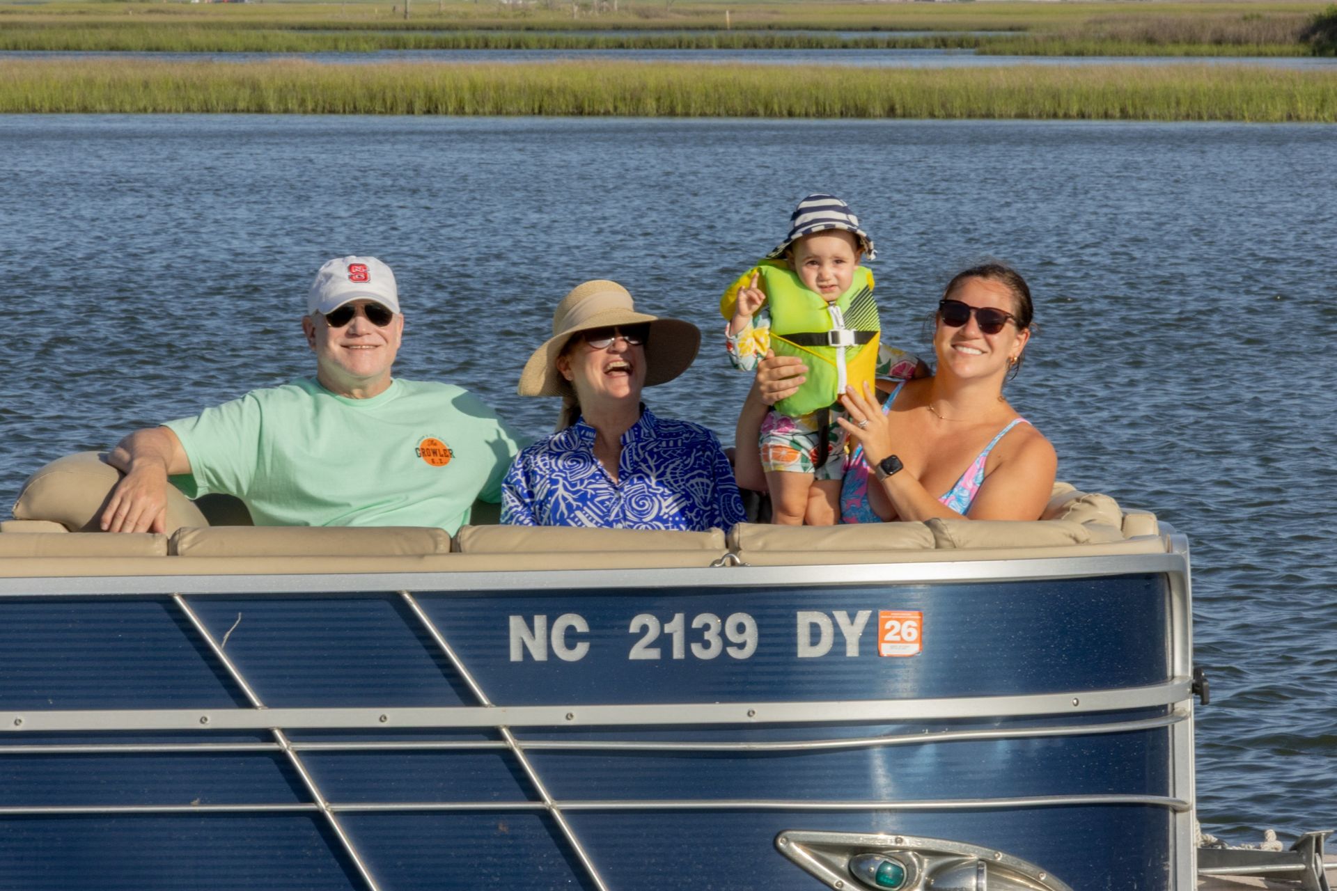 Four people smiling on a pontoon boat on a lake, with blue water and grassy shoreline behind them.