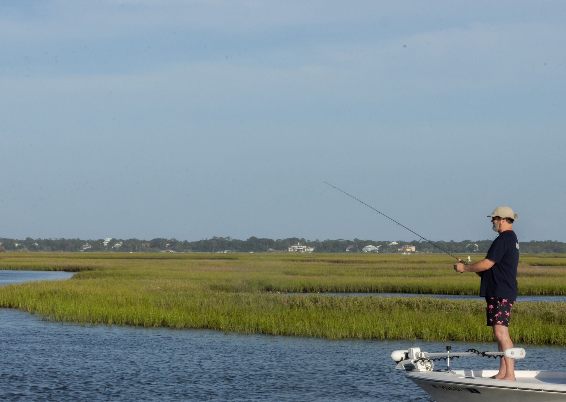 Person fishing from a small boat on calm water beside grassy marshland under a clear sky.