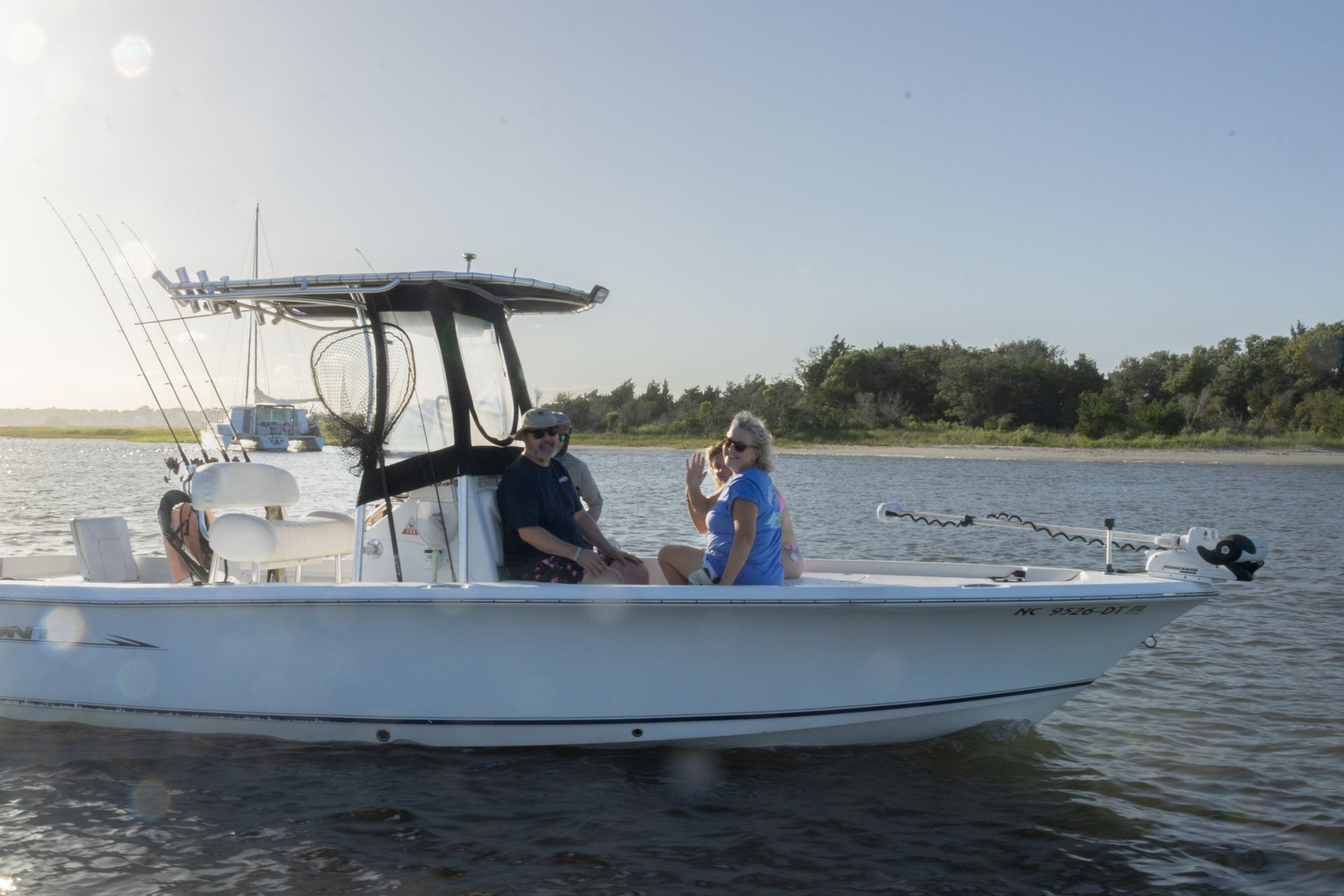 Two people on a small white motorboat on calm water near a wooded shoreline