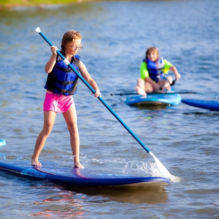 Two children paddle standing on blue paddleboards in shallow water, one in foreground with a blue paddle