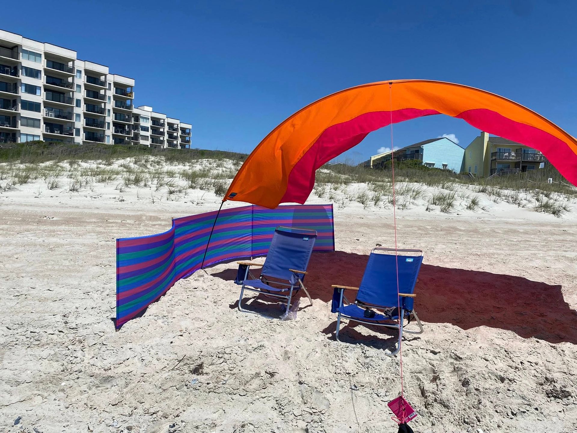 Beach setup with two blue chairs, a striped windbreak, and an orange canopy on a sunny sandy shore