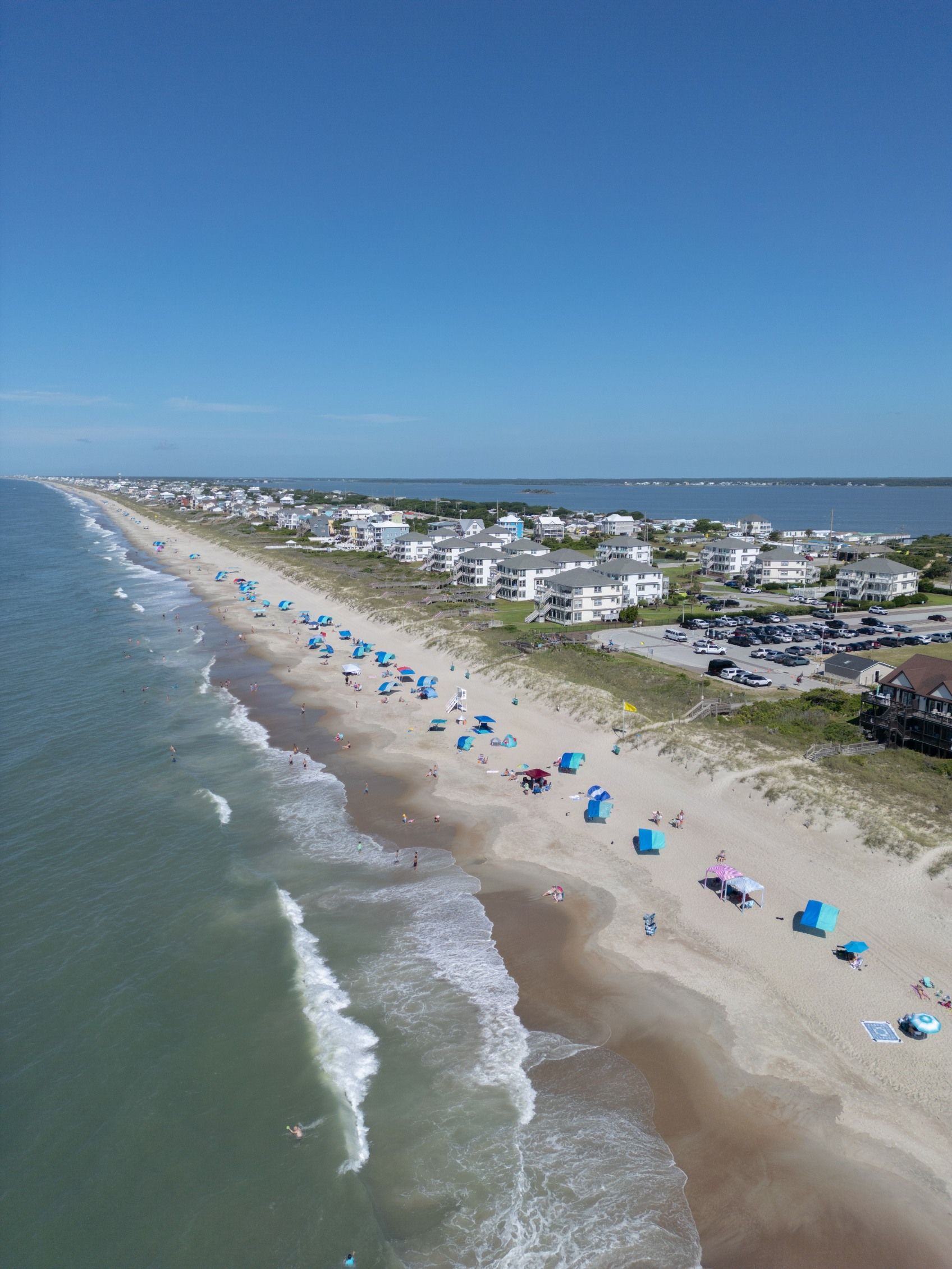 Aerial view of a sandy beach with blue umbrellas, ocean waves, and seaside houses under a clear sky
