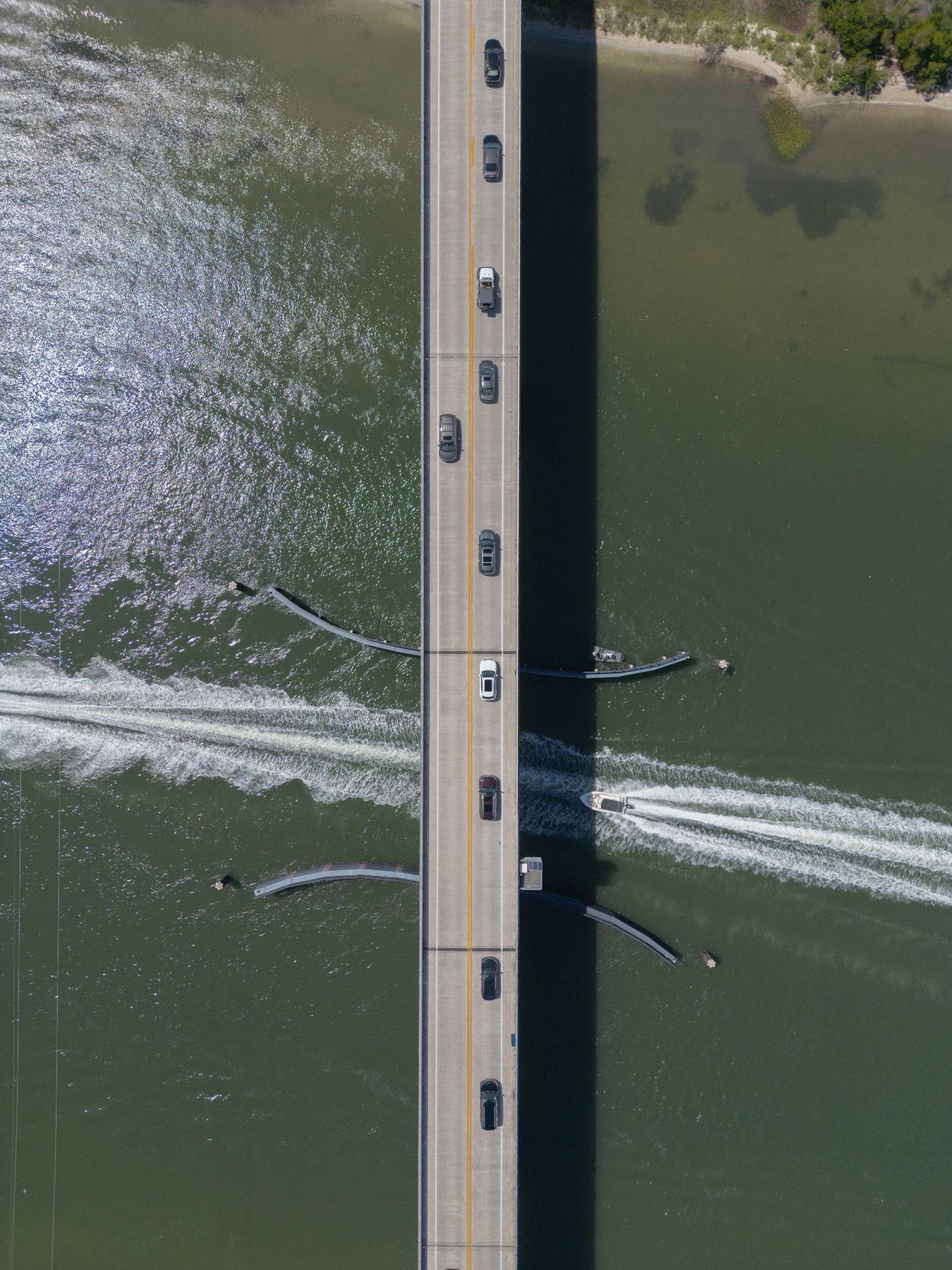 Aerial view of a bridge over green water with several boats leaving wakes on both sides.
