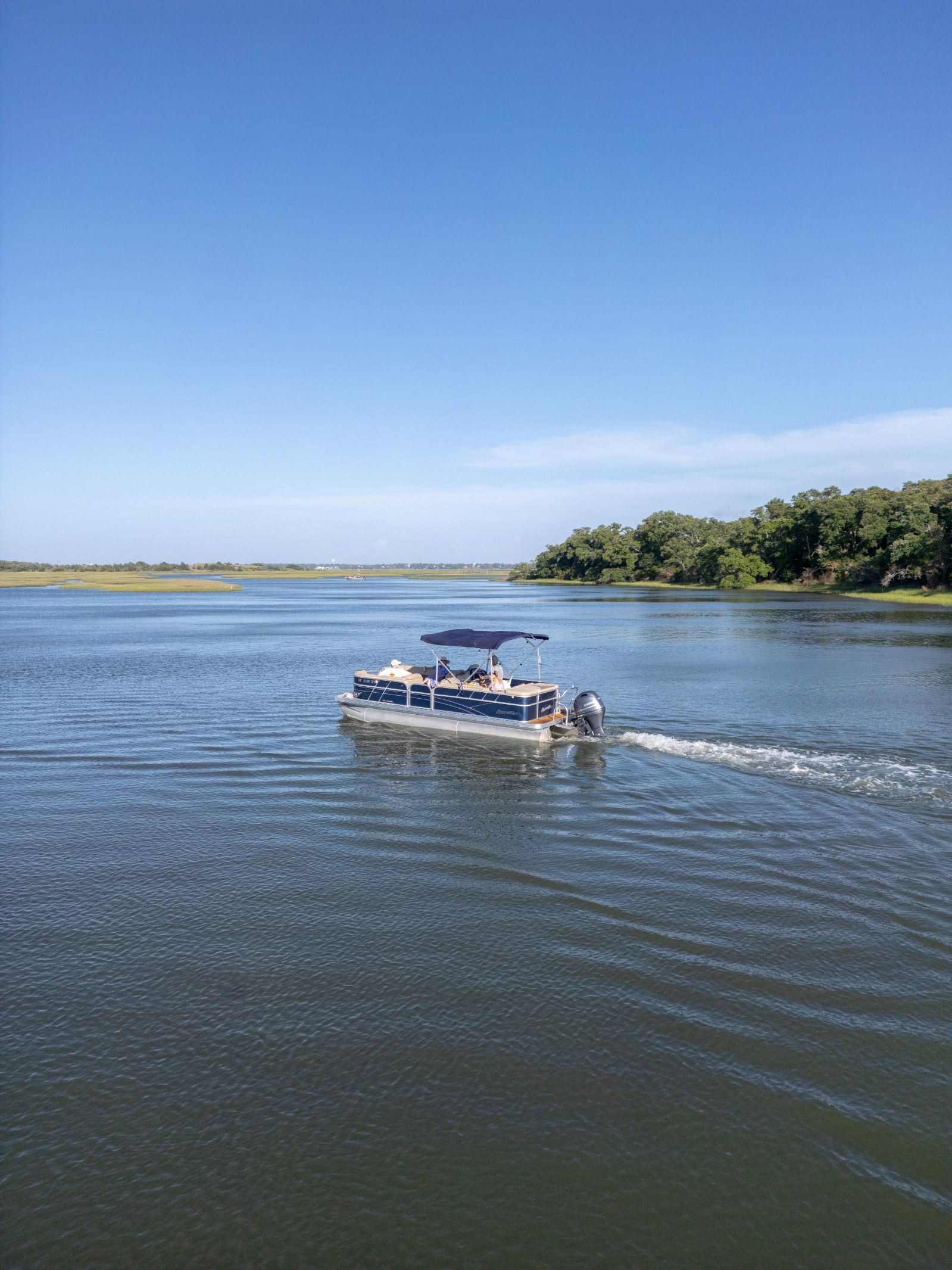 Pontoon boat cruising on a calm river under a clear blue sky, with trees along the shore