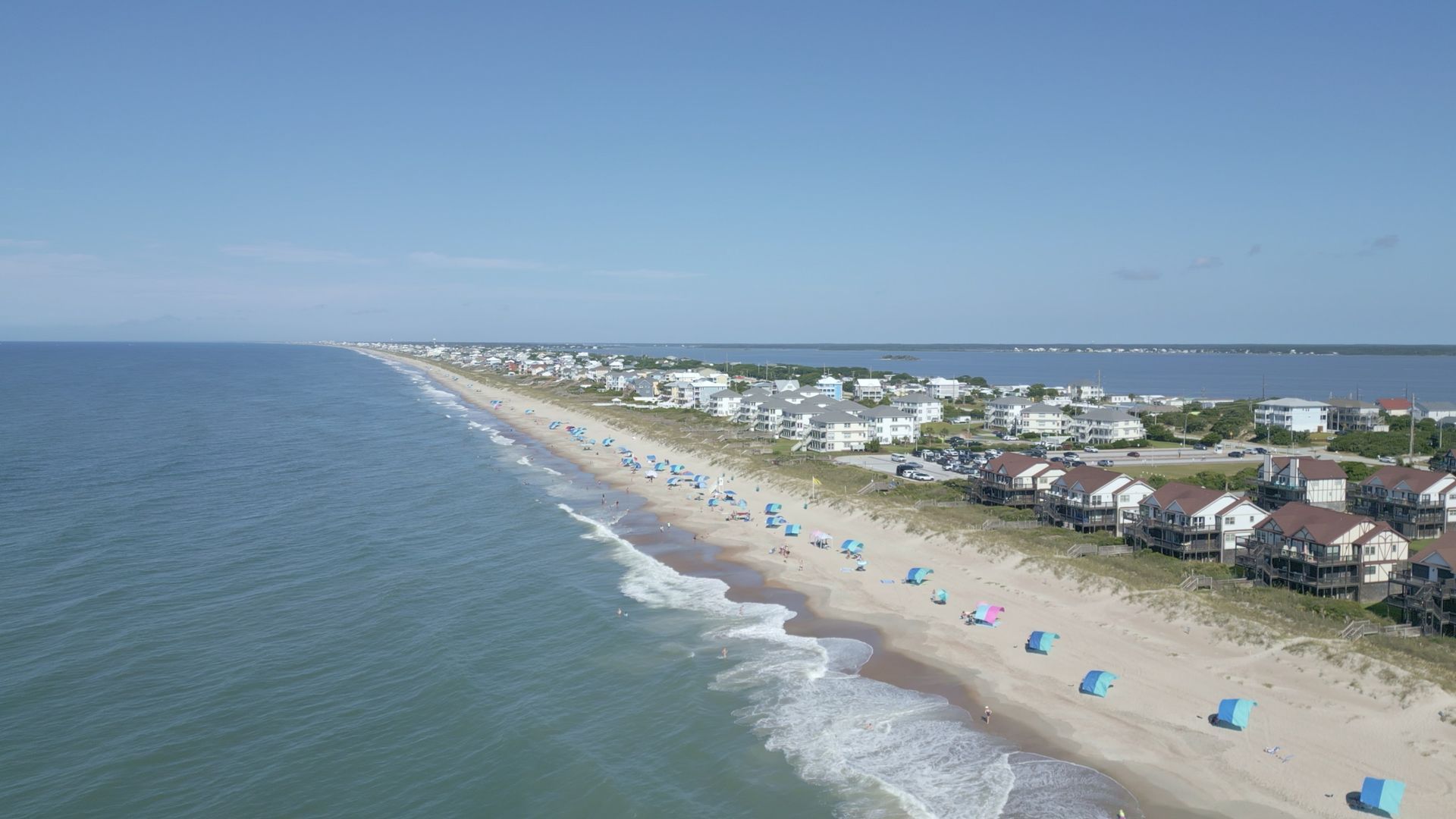 Aerial view of a sandy beach with blue umbrellas, ocean waves, and houses along the shoreline.
