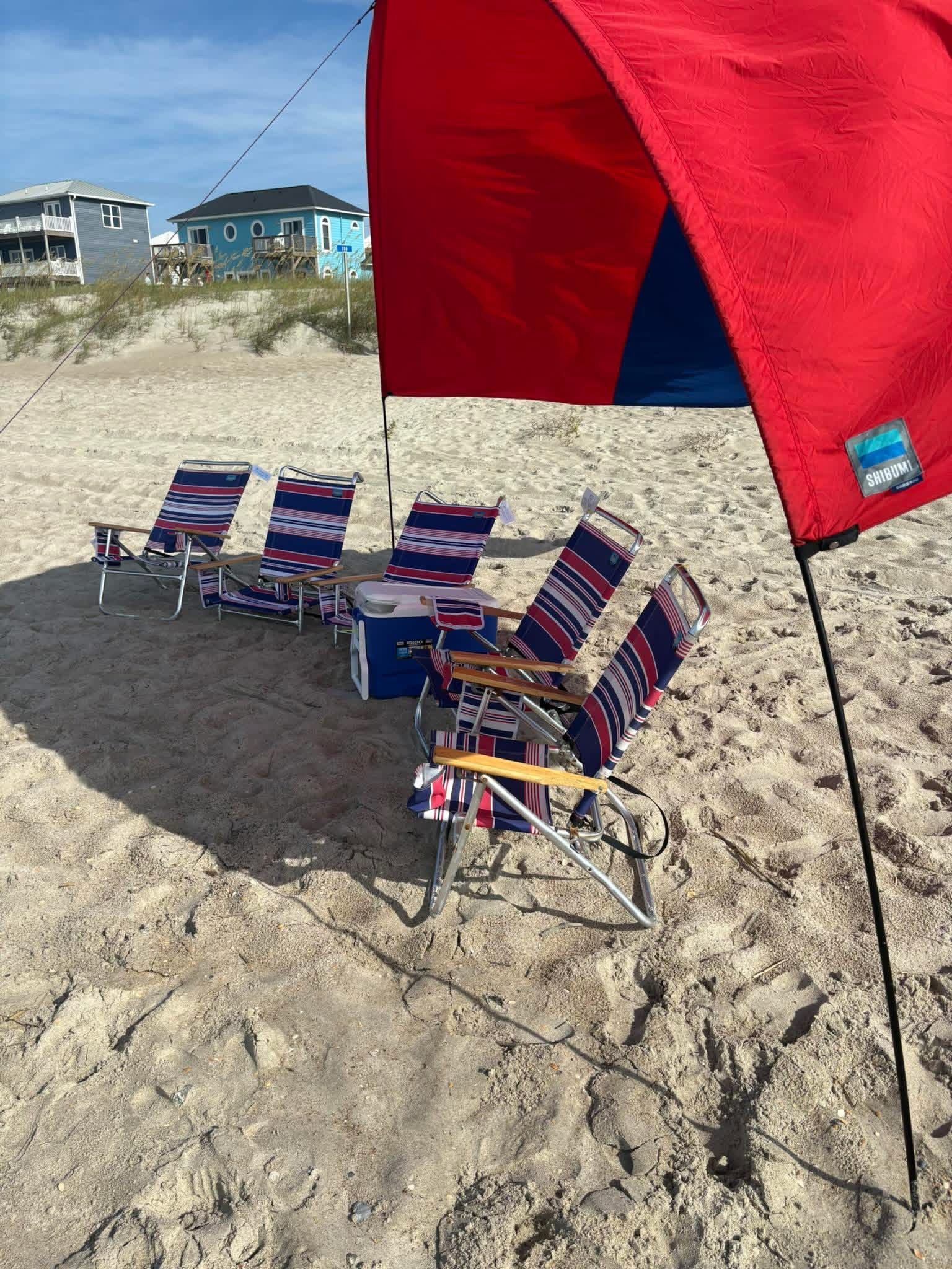 Red beach canopy shading striped folding chairs on a sandy beach near oceanfront houses.