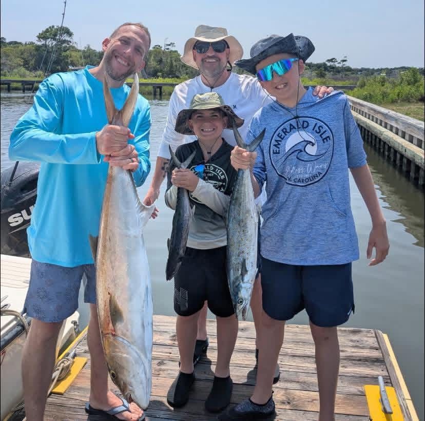Four people on a dock hold up a large fish in bright daylight.