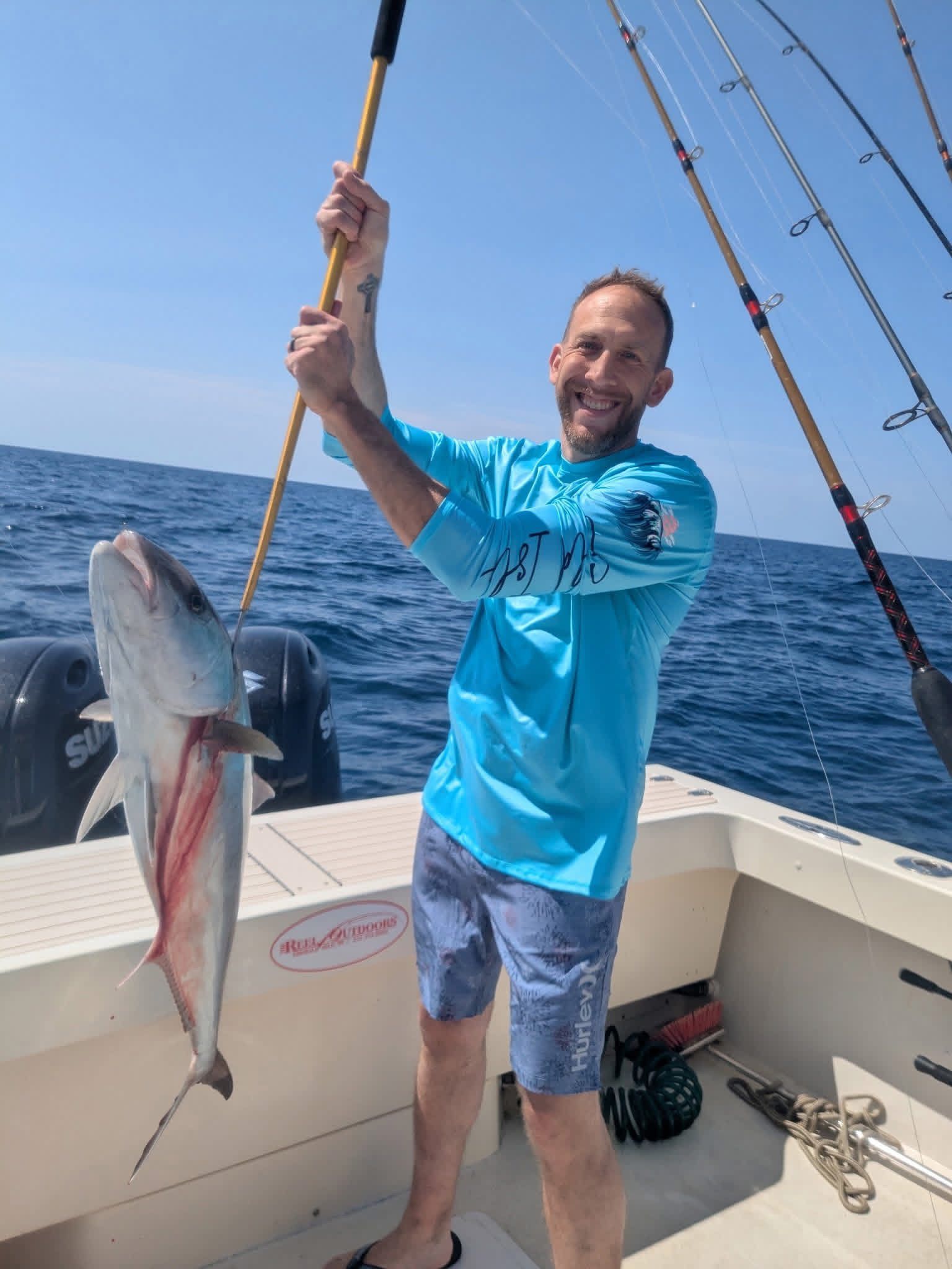 Man holding a large fish on a boat at sea, smiling and wearing a blue shirt.