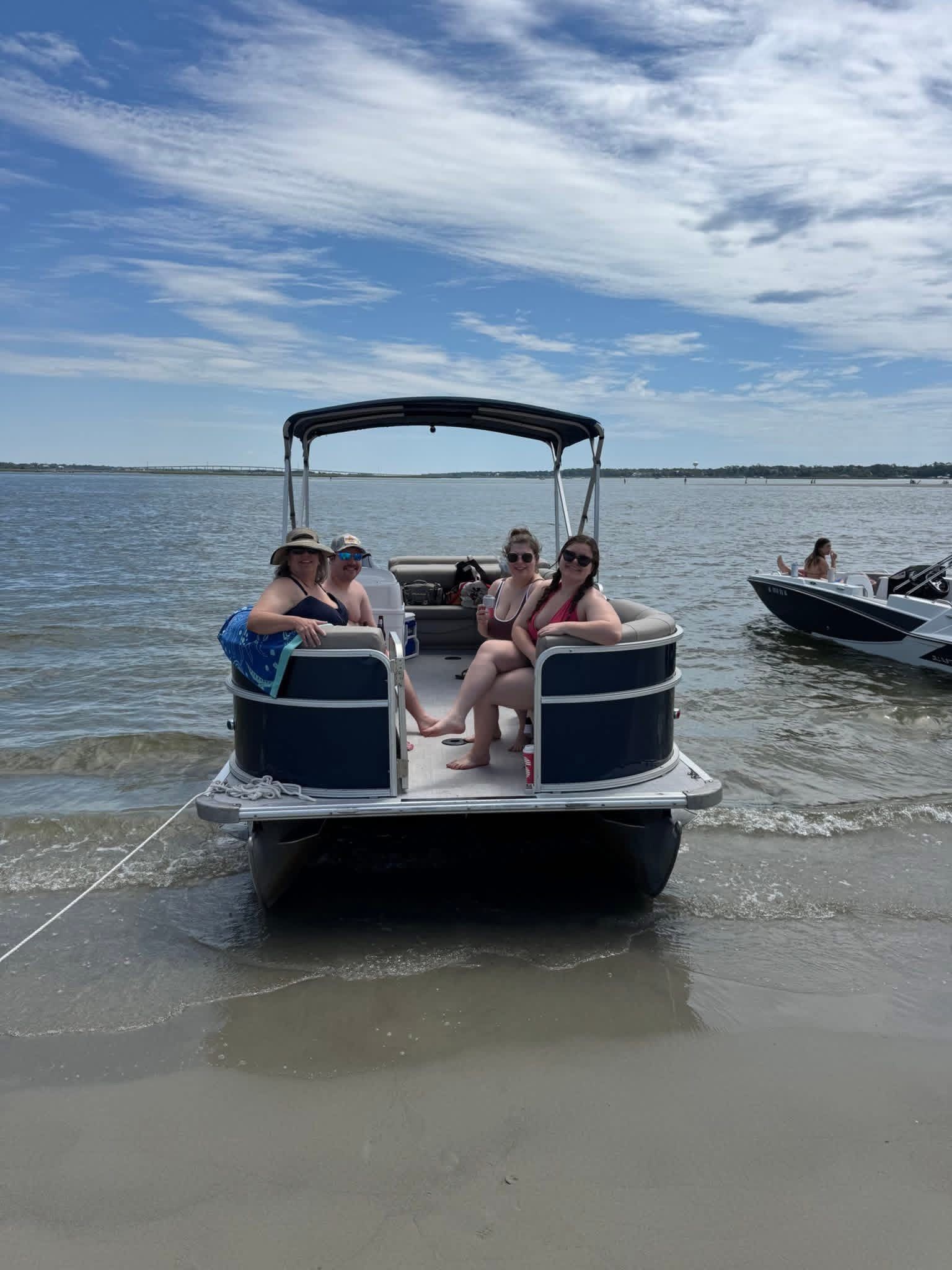 Pontoon boat with people boarding from the beach, floating on calm water under a partly cloudy sky
