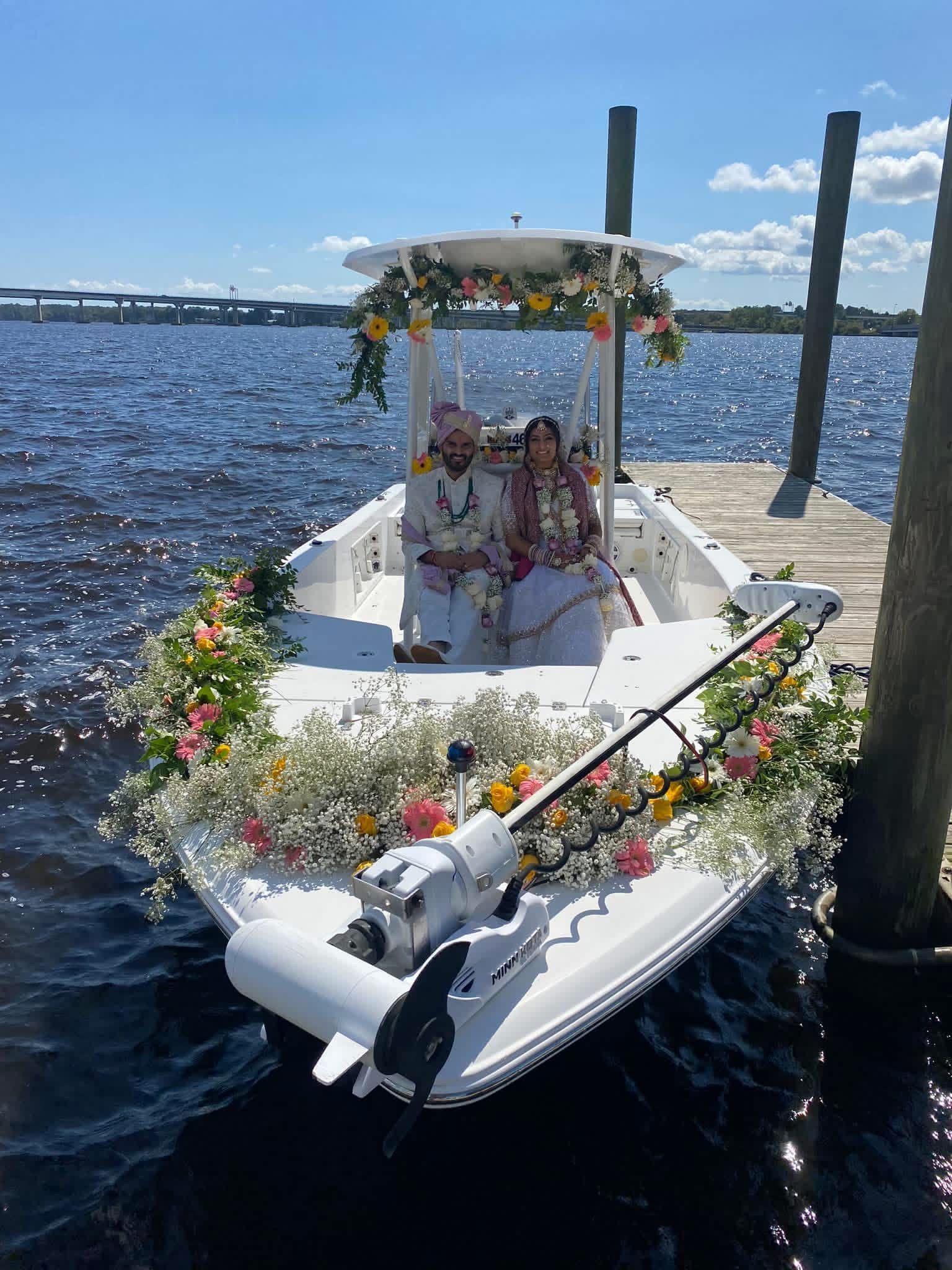 Decorated white boat docked at a pier on a sunny lake, with people seated under a floral canopy.