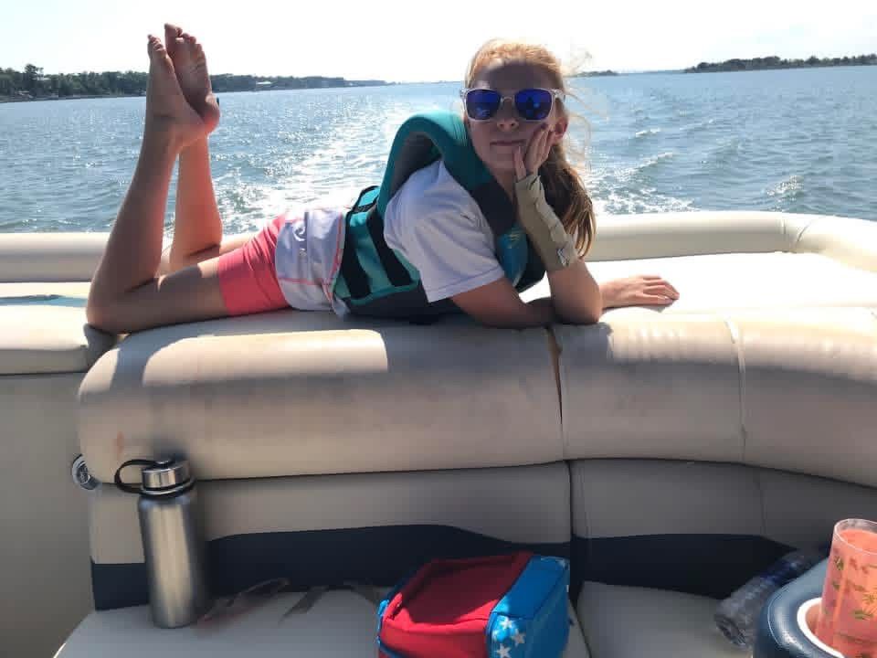 Person lounging on a boat seat, smiling in sunglasses on a sunny lake with a wake behind.