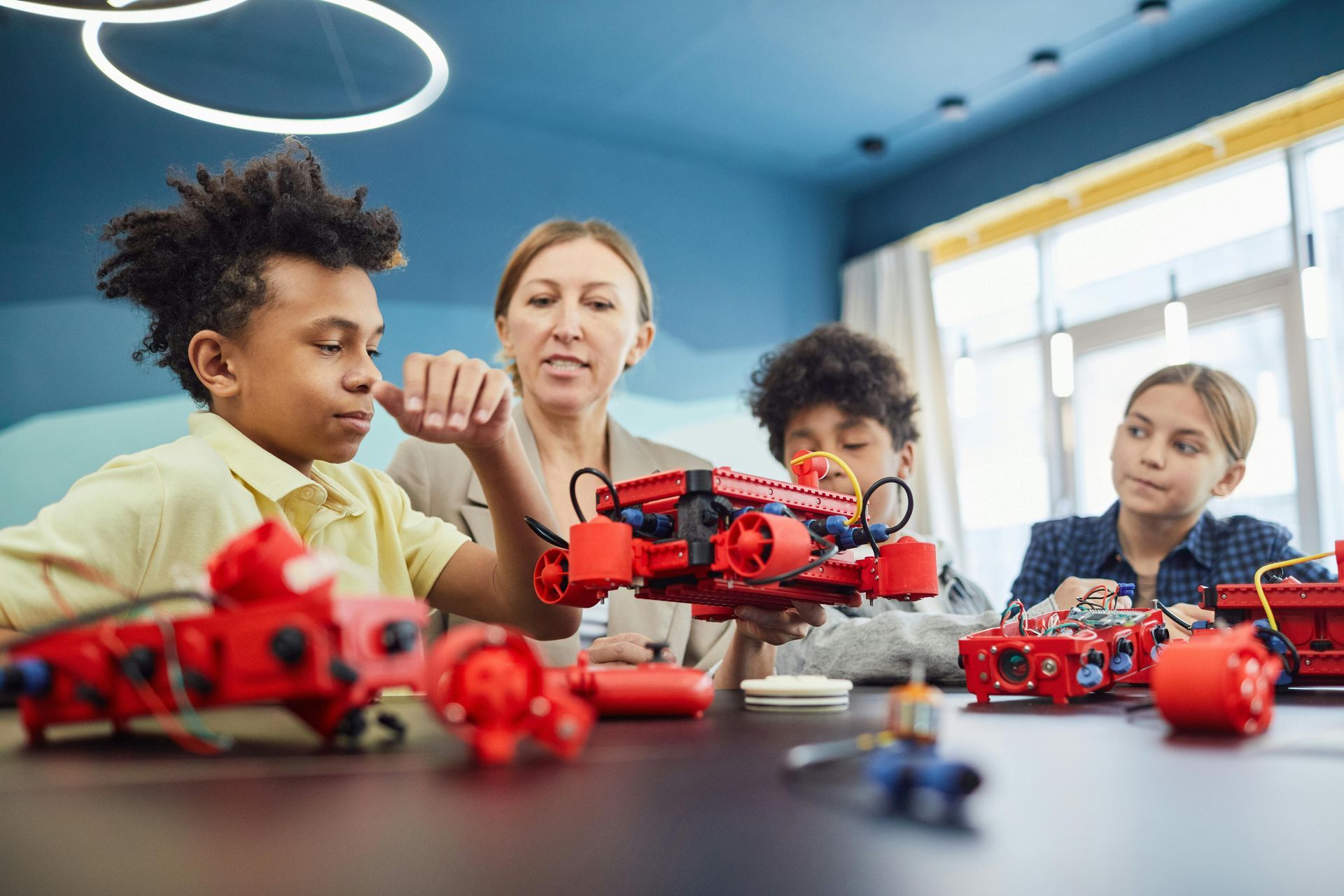 Children and an adult gather around red robot cars in a bright classroom, focusing on a robotics project.
