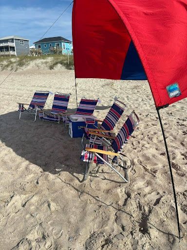 Beach chairs under a red canopy on sandy dunes, with houses in the background.